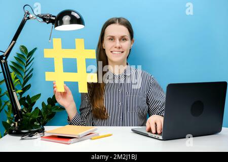 Jeune employée souriante jeune femme assise sur le lieu de travail avec un ordinateur portable et montrant un grand symbole jaune hashtag, partageant du contenu viral, message balisé, posi Banque D'Images
