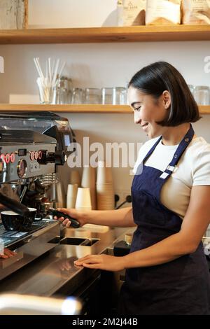 Bonne fille asiatique, le barista en tablier travaille avec une machine à café derrière le comptoir, debout dans un café et prépare du cappuccino Banque D'Images