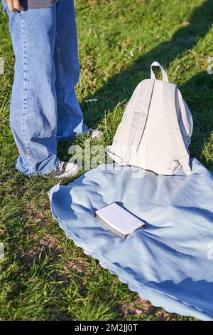 Photo verticale, corps de jeune fille adolescente, debout à côté d'une couverture avec sac à dos et livre, pique-nique à l'extérieur Banque D'Images