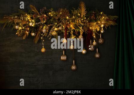 Une couronne d'arbre de Noël avec guirlandes et jouets est accrochée sur un mur vert à côté d'un rideau vert, décor du nouvel an Banque D'Images