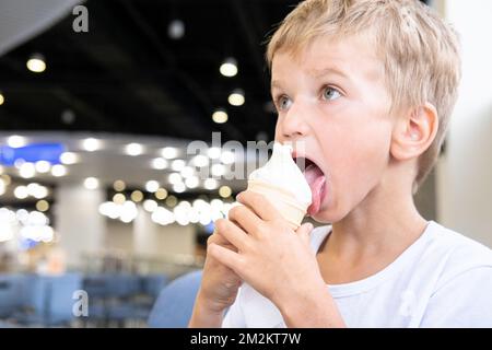 Portrait d'un petit garçon drôle et affamé qui mange de la glace froide délicieuse dans une tasse de gaufres assis dans un café, fond sombre, bokeh. Un enfant aime la glace Banque D'Images