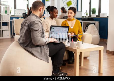 Homme d'affaires tenant un ordinateur portable avec des professionnels de la finance, tandis que divers employés parlent d'expliquer le plan de projet en examinant les documents administratifs assis sur le canapé. Collègues multiethniques analysant les rapports financiers Banque D'Images