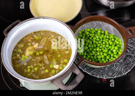 Préparation de soupe aux pois, bouillon dans une casserole sur une table de cuisson en céramique chaude, pois dans une passoire, couvercle. Banque D'Images