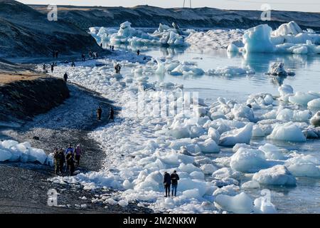 On voit des gens marcher près des zones de glace sur la rive du lac Jökulsárlón. Jökulsárlón est un lac situé dans le sud de l'Islande, avec une superficie de 20 km2 et une profondeur de plus de 200 mètres. Jusqu'à il y a moins de 100 ans, le glacier de Breiðamerkurjökull (qui fait partie du glacier Vatnajökull) s'étendait même au-delà du périphérique. En raison de la hausse des températures, le glacier a reculé, créant cette impressionnante lagune glaciaire. Banque D'Images