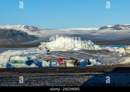 On voit des gens marcher près des zones de glace sur la rive du lac Jökulsárlón. Jökulsárlón est un lac situé dans le sud de l'Islande, avec une superficie de 20 km2 et une profondeur de plus de 200 mètres. Jusqu'à il y a moins de 100 ans, le glacier de Breiðamerkurjökull (qui fait partie du glacier Vatnajökull) s'étendait même au-delà du périphérique. En raison de la hausse des températures, le glacier a reculé, créant cette impressionnante lagune glaciaire. Banque D'Images