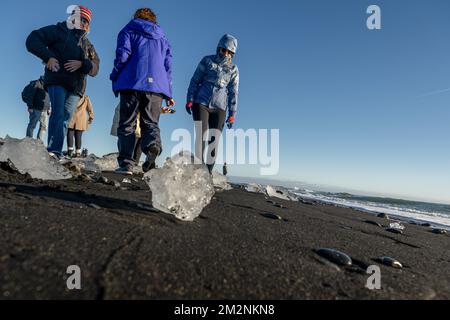 On voit des gens marcher autour des blocs de glace sur la plage de Diamond, près de Jökulsárlón. Jökulsárlón est un lac situé dans le sud de l'Islande, avec une superficie de 20 km2 et une profondeur de plus de 200 mètres. Jusqu'à il y a moins de 100 ans, le glacier de Breiðamerkurjökull (qui fait partie du glacier Vatnajökull) s'étendait même au-delà du périphérique. En raison de la hausse des températures, le glacier a reculé, créant cette impressionnante lagune glaciaire. Banque D'Images