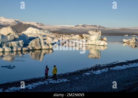 On voit des gens marcher près des zones de glace sur la rive du lac Jökulsárlón. Jökulsárlón est un lac situé dans le sud de l'Islande, avec une superficie de 20 km2 et une profondeur de plus de 200 mètres. Jusqu'à il y a moins de 100 ans, le glacier de Breiðamerkurjökull (qui fait partie du glacier Vatnajökull) s'étendait même au-delà du périphérique. En raison de la hausse des températures, le glacier a reculé, créant cette impressionnante lagune glaciaire. Banque D'Images