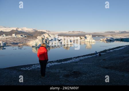 On voit une personne marcher près de la rive du lac Jökulsárlón. Jökulsárlón est un lac situé dans le sud de l'Islande, avec une superficie de 20 km2 et une profondeur de plus de 200 mètres. Jusqu'à il y a moins de 100 ans, le glacier de Breiðamerkurjökull (qui fait partie du glacier Vatnajökull) s'étendait même au-delà du périphérique. En raison de la hausse des températures, le glacier a reculé, créant cette impressionnante lagune glaciaire. Banque D'Images