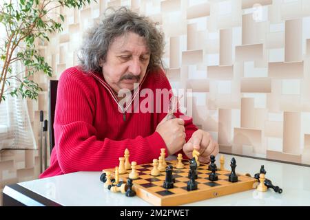 Un retraité dans un chandail rouge et des lunettes dans sa main assis à la table et jouant aux échecs Banque D'Images