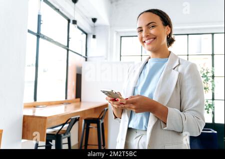 Photo d'une femme hispanique ou brésilienne positive, entrepreneur d'affaires, debout dans un bureau moderne, utilise son téléphone mobile, messagerie en ligne avec un collègue ou un client, regarde la caméra, souriant amical Banque D'Images
