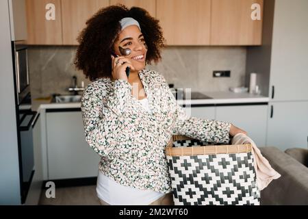 Jeune belle femme africaine souriante tenant un panier de salon et un téléphone parlant à la maison Banque D'Images