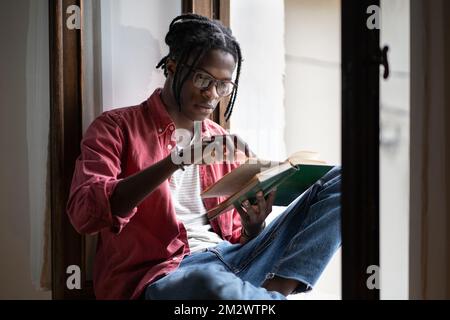 Un homme afro-américain concentré et sans émotion dans une tenue décontractée lisant le livre préféré se trouve sur le seuil de la fenêtre Banque D'Images