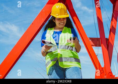 jeune femme latine caucasienne ingénieur dans une construction à l'extérieur, elle sourit de lire des messages et des notifications sur son téléphone, en utilisant des applications mobiles et Banque D'Images