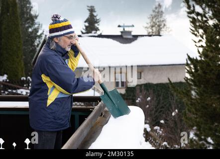 Un homme adulte aux cheveux gris se tient sur le balcon et nettoie la neige de la rampe avec une pelle Banque D'Images