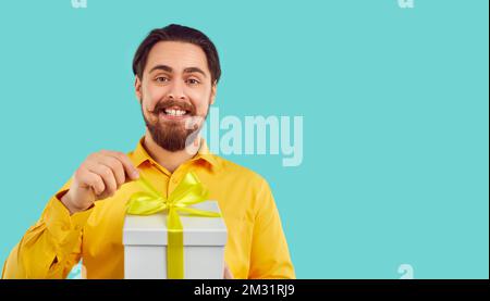 Homme excité avec expression heureuse unties noeud sur boîte cadeau isolé sur fond bleu clair. Banque D'Images