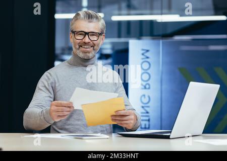 Heureux homme senior assis dans le bureau à la table et tenant une enveloppe dans sa poche. J'ai reçu de bonnes nouvelles, un prêt. J'ai fait un contrat. Il regarde la caméra, sourit. Banque D'Images