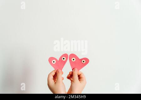 Fait à la main pour la Saint-Valentin ou la fête des mères. Deux coeurs en papier rose peints à la main avec les yeux dans les mains de l'enfant. Arrière-plan blanc. Copier l'espace Banque D'Images