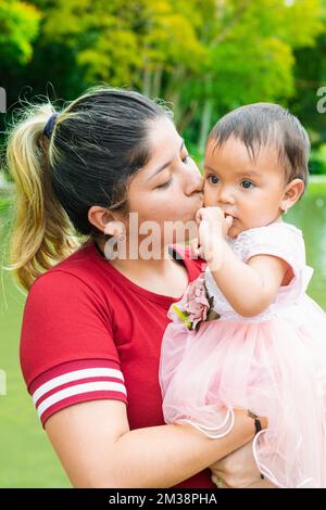 latina mère embrasse son petit bébé brunette tout en la tenant dans ses bras, bébé distrait tout en étant embrassé sur sa joue Banque D'Images
