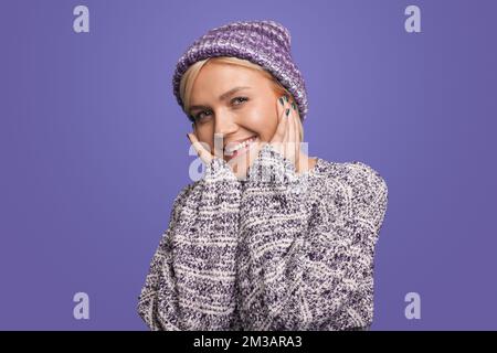 Portrait d'une belle femme blonde portant un chandail et un chapeau en laine, regardant l'appareil photo isolé sur fond violet. Concept de bonheur. Positif Banque D'Images