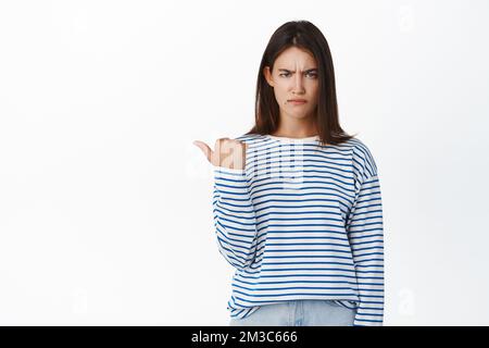 Une fille de brunette en colère pointant vers la gauche, regardant avec désapprobation et l'expression du visage de pouting bouleversé, debout dans un chandail sur fond blanc Banque D'Images