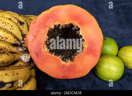 Vue rapprochée d'une papaye d'orange fraîchement coupée en tranches avec graines à l'intérieur, d'un groupe de bananes freckled et de trois citrons verts sur un backgroun bleu marine Banque D'Images