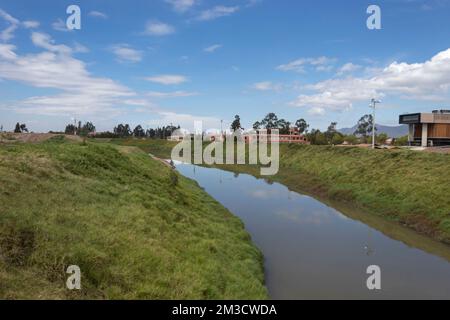 La rivière Bogota traverse un champ de campagne vert avec des bâtiments modernes à côté et un ciel bleu à l'arrière-plan en journée ensoleillée Banque D'Images