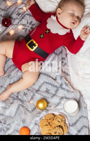 Enfant mignon dormant dans le costume du Père Noël à côté du lait et des biscuits debout Banque D'Images