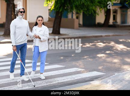 Jeune femme aidant un homme aveugle senior à traverser la route en ville Banque D'Images