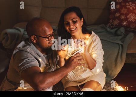 Heureux couple interracial célébrant le nouvel an à la maison ensemble, assis près de l'arbre de Noël Banque D'Images