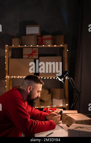 bonnes recettes pendant la vente de noël. Un jeune homme d'affaires indien barbu est assis à une table la nuit dans un costume rouge vérifie les commandes sur le site usi Banque D'Images