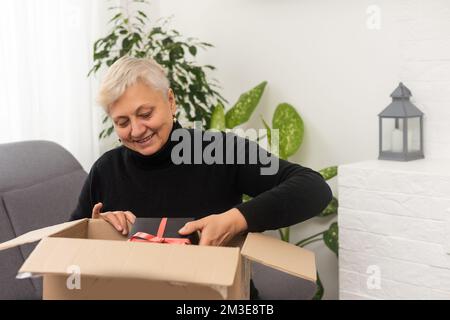 Une femme d'âge moyen positive assise sur un canapé avec une boîte en carton sur des tours, se sentant curieuse de commander l'article souhaité sur le site Web, souriant femme Banque D'Images