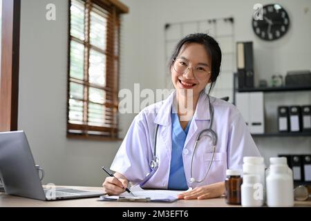 Belle femme asiatique millénaire médecin en uniforme s'assoit à son bureau, souriant et regardant la caméra. Banque D'Images