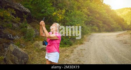 Bonne femme touriste senior marchant sur la route forestière d'été à l'extérieur à l'heure du coucher du soleil. Une vieille femme mince voyageant avec un appareil photo fait le selfie sur un smartphone. Concept de vacances de retraite actives. Filtre chaud Banque D'Images