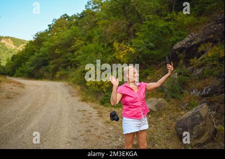 Bonne femme touriste senior marchant sur la route forestière d'été à l'extérieur à l'heure du coucher du soleil. Une vieille femme mince voyageant avec un appareil photo fait le selfie sur un smartphone. Concept de vacances de retraite actives. Filtre chaud Banque D'Images