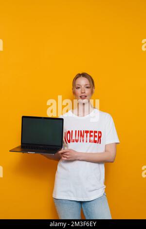 Jeune femme blonde portant un t-shirt montrant un ordinateur portable isolé sur fond jaune Banque D'Images