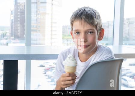 Un jeune garçon mignon mange une glace blanche tourbillonnante dans une tasse à gaufres, assis à une table près de la fenêtre dans un café. Arrière-plan flou. Jour d'été. Le CH Banque D'Images