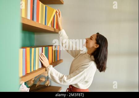 Belle jeune femme prenant le livre de l'étagère dans la bibliothèque à la maison Banque D'Images