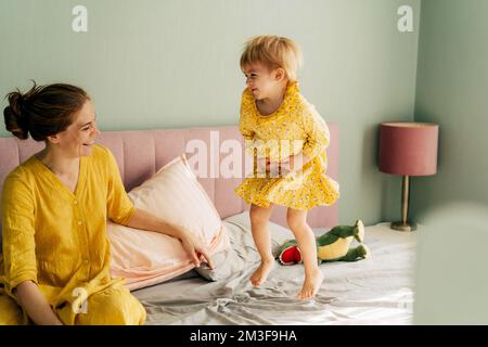 Une petite fille saute gaiement sur le lit, maman rit et s'assoit à côté d'elle. Banque D'Images