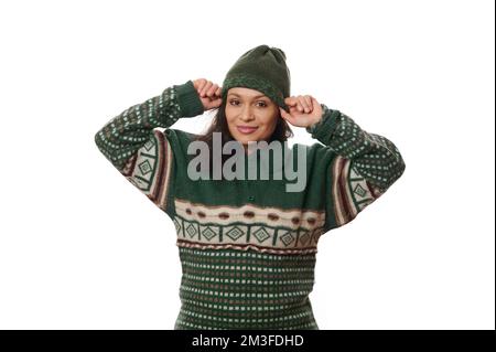 Femme brunette multi-ethnique, dans un chandail vert chaud, mettant un chapeau de laine sur sa tête, exprimant des émotions positives Banque D'Images