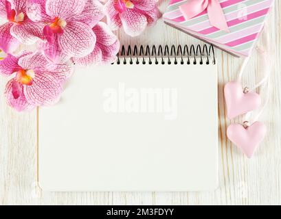 Arrière-plan de la Saint-Valentin avec bloc-notes vide pour le texte, boîte-cadeau, fleur d'orchidée rose et coeurs sur une surface en bois blanc. Plan d'agencement, vue de dessus. Banque D'Images