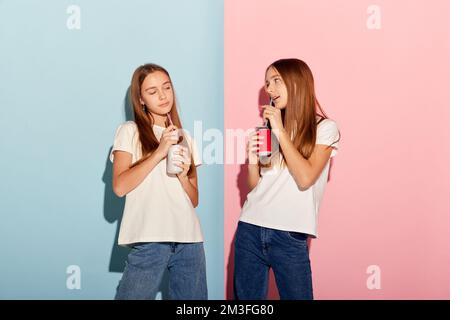 Deux belles filles adolescentes, frères et sœurs en t-shirts et jeans dégustation de limonade isolée sur fond duotone rose-bleu. Émotions, famille, plaisir, joie Banque D'Images