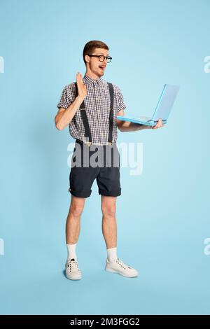 Portrait d'un jeune homme en chemise à carreaux, short et bretelles ayant un appel vidéo sur ordinateur portable isolé sur fond bleu. Réunion Internet Banque D'Images