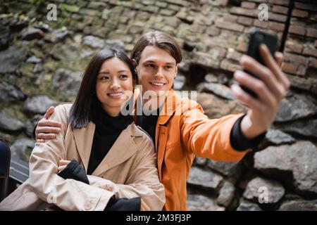 Homme souriant embrassant petite amie asiatique tout en prenant selfie sur smartphone sur la terrasse du café, image de stock Banque D'Images