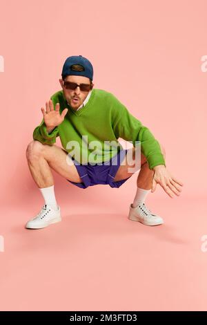 Portrait d'un jeune homme dans des vêtements à l'ancienne posant, assis sur des squats isolés sur fond rose. Swag Banque D'Images