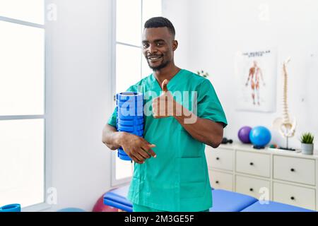 Un jeune physiothérapeute africain tenant un rouleau en mousse à la clinique souriant heureux et positif, pouce vers le haut faisant excellent et signe d'approbation Banque D'Images