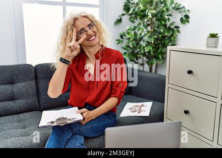 Femme psychologue d'âge moyen au bureau de consultation faisant le symbole de la paix avec les doigts sur le visage, souriant gai montrant la victoire Banque D'Images
