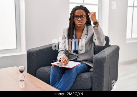 Un jeune afro-américain aux tresses qui travaillent au bureau de consultation est en colère et fou pour soulever un poing frustré et furieux tout en criant avec colère. enrager un Banque D'Images