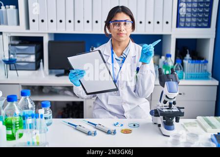 Jeune femme hispanique travaillant au laboratoire scientifique pointant du doigt vers le côté montrant la publicité, sérieux et calme visage Banque D'Images