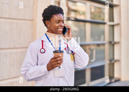Femme afro-américaine portant l'uniforme du médecin parlant sur le smartphone en train de boire du café dans la rue Banque D'Images