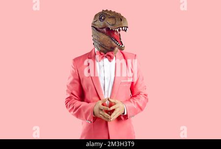 Homme drôle dans un masque de dinosaure en caoutchouc et un costume rose debout isolé sur un fond rose Banque D'Images